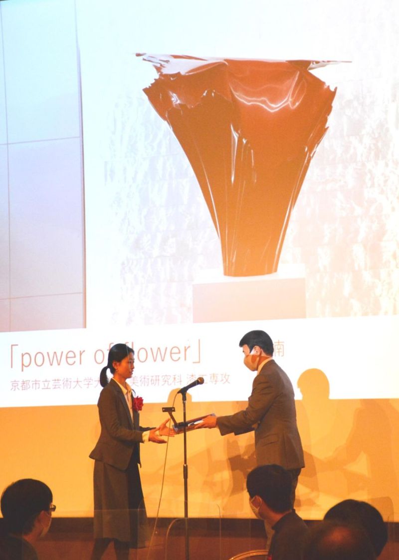 服部社長(右)から表彰を受ける隗さん。スクリーンは最優秀賞受賞作品